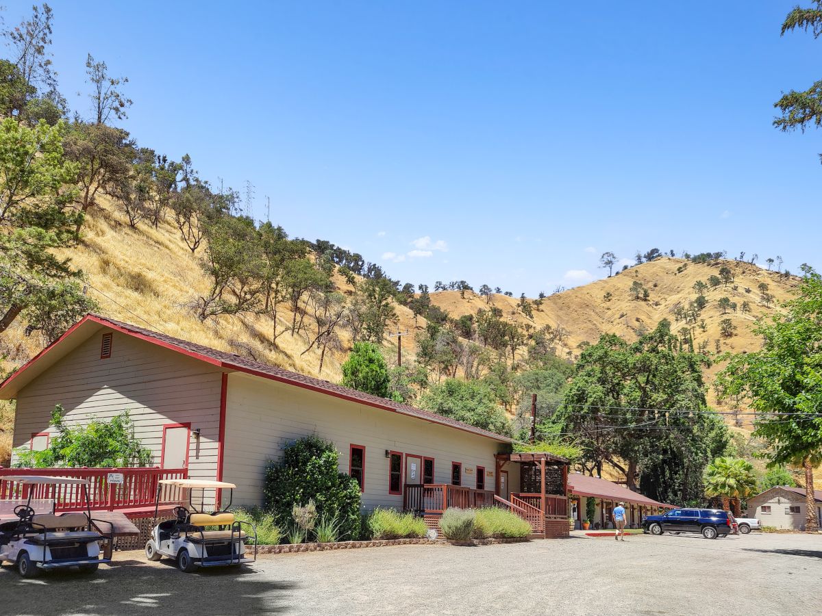 A small, single-story motel or lodge with red trim sits beside a dirt lot, set against rolling, tree-dotted hills under a clear blue sky.
