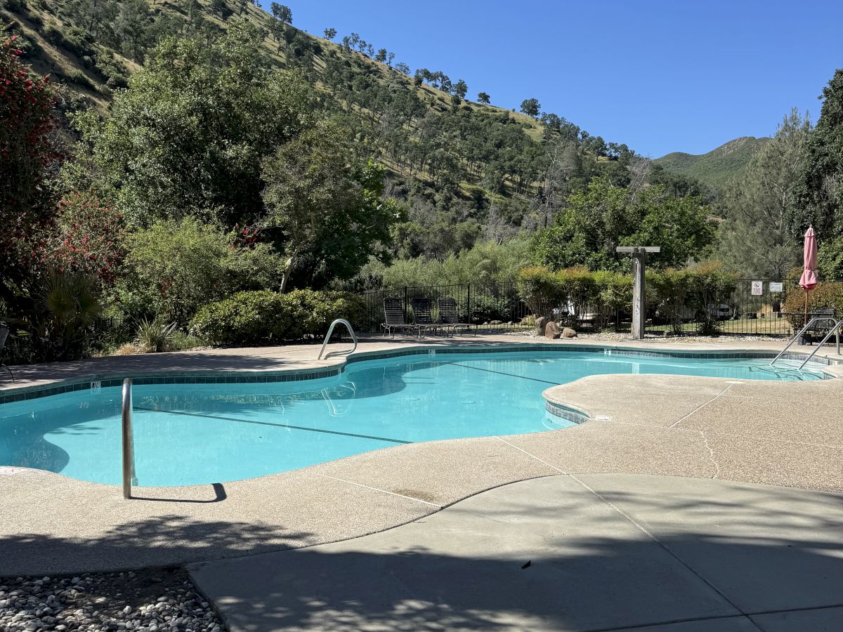 A sunny outdoor pool area surrounded by hills and trees, with clear blue water, deck, and a few structures in the background.