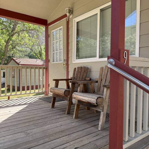 A shaded porch with two wooden Adirondack chairs, a railing, and a door with a window; trees in the background and sunny weather.