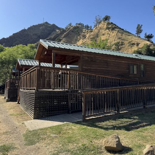Wooden cabins with green roofs sit along a railinged porch, set against grassy ground and hills under a clear blue sky.
