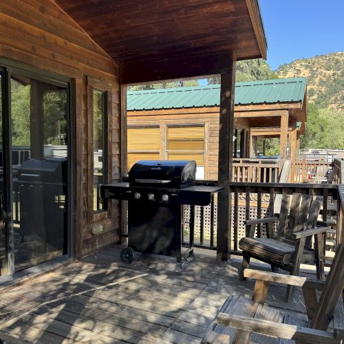 A wooden deck with a barbecue grill and chairs, attached to a cabin, under a shaded overhang, with mountains in the background.
