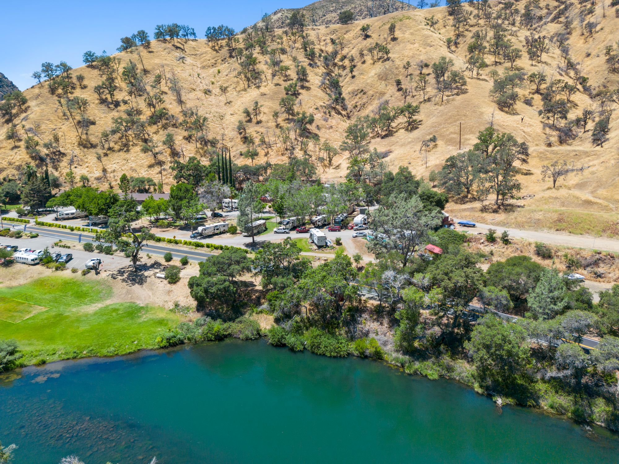 An aerial view shows a lakeside park with a grassy field, trees lining the water, a row of houses or cabins near a road, and a brown hillside in the background.