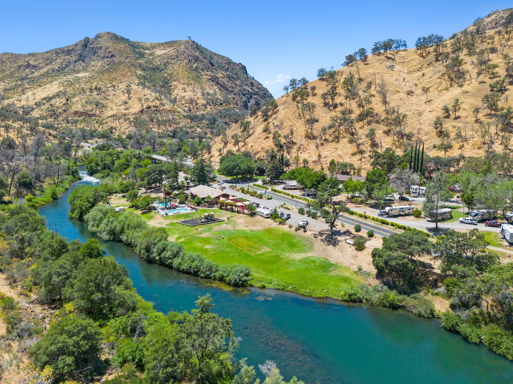 A scenic river winds through a green park by canyon walls, with a small village, trees, and blue skies in a sunny valley.