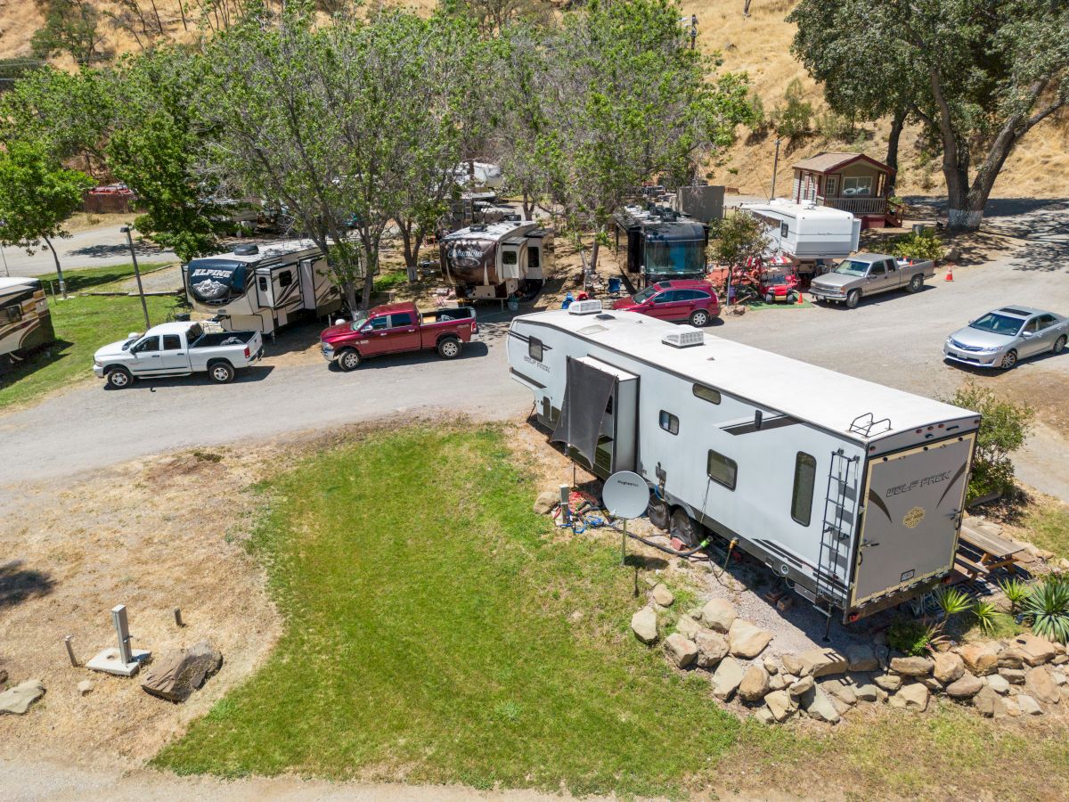 Aerial view of a campground with a large RV, several parked cars, and trees; a grassy area and picnic stones surround the trailer site.